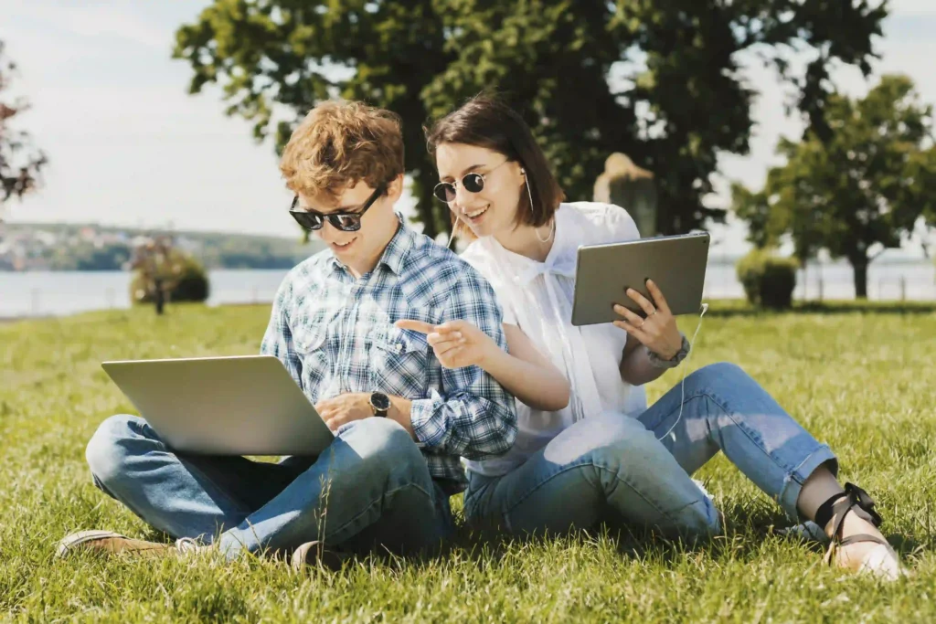 Zwei junge Menschen sitzen auf einer Wiese und arbeiten gemeinsam mit einem Laptop und einem Tablet. Die beiden tragen Sonnenbrillen und lachen. Im Hintergrund sind Bäume und ein See zu sehen.