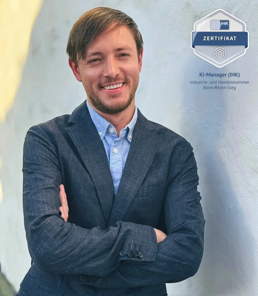 Porträt eines professionellen Mannes in einem dunkelblauen Blazer und einem hellblauen Hemd, der vor einer strukturierten weißen Wand steht. Er hat helle Haut, einen kurzen Bart und braunes Haar, das zur Seite gekämmt ist. Der Mann hat einen freundlichen Ausdruck, mit einem sanften Lächeln und entspannter Haltung, die Arme vor der Brust verschränkt. Auf der rechten Seite ragt ein IHK-Zertifikat als KI-Manager der Industrie- und Handelskammer Bonn/Rhein-Sieg hervor.